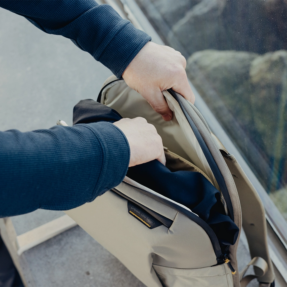 Person opening a small beige backpack with a blue garment inside, viewed from the side.