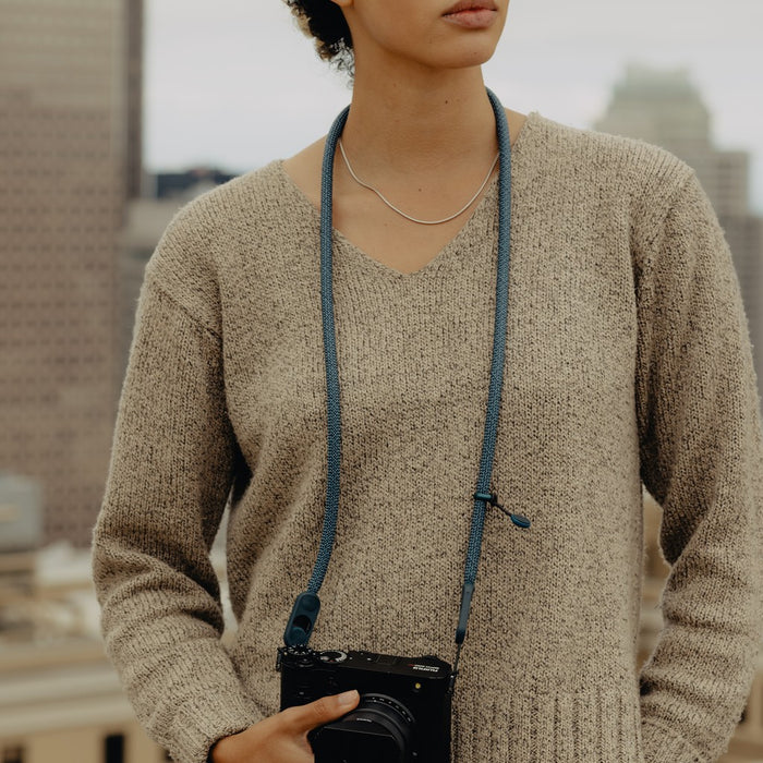 Person wearing a beige sweater holding a camera with a blue strap.