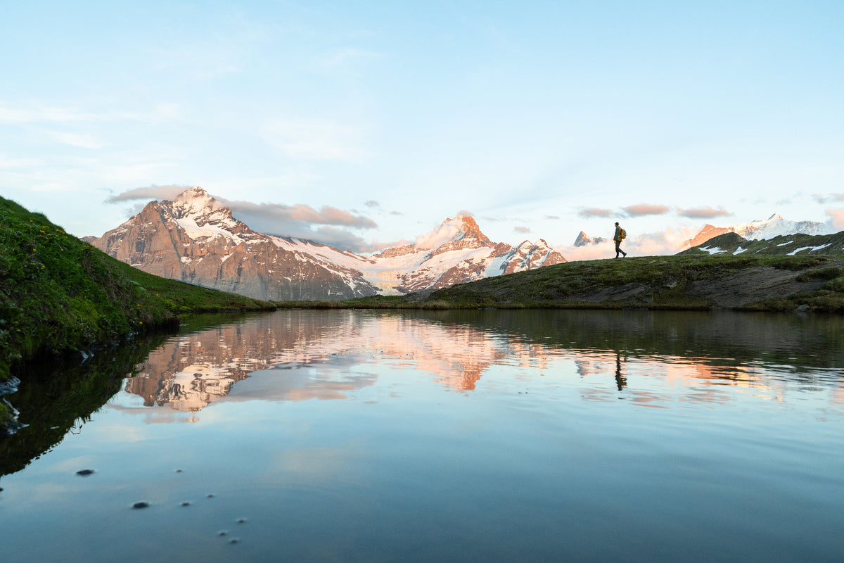 A scenic landscape photograph of snow-capped mountains reflected in a calm lake, with a small hiker standing on a grassy ridge under a soft blue sky.
