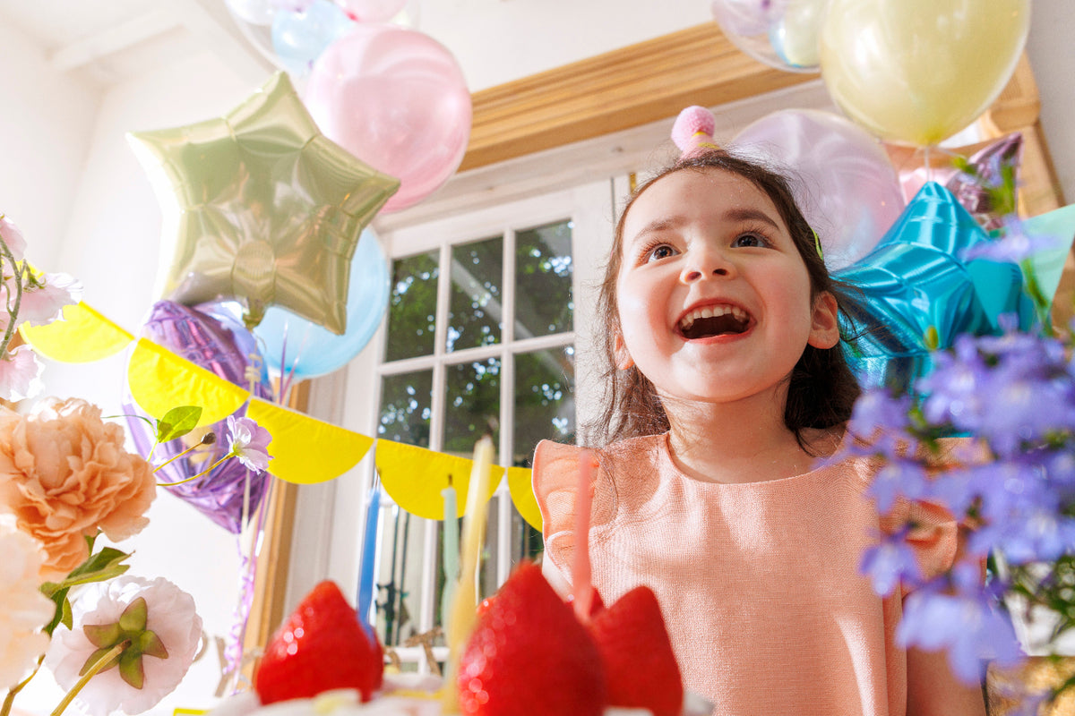 A young girl with a pink party hat smiles joyfully at a birthday celebration, surrounded by colorful balloons, star-shaped foil decorations, flowers, and a strawberry cake in a brightly lit room.