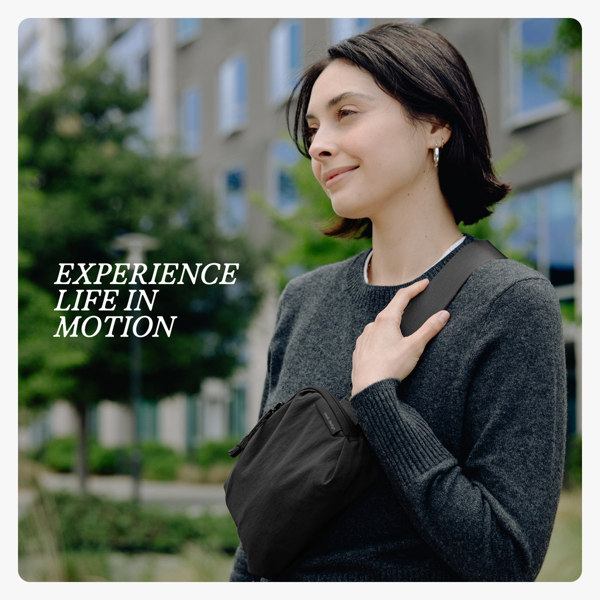 A woman wearing a dark gray knit sweater and carrying a black crossbody bag outdoors, with the text 'EXPERIENCE LIFE IN MOTION' on the left side.