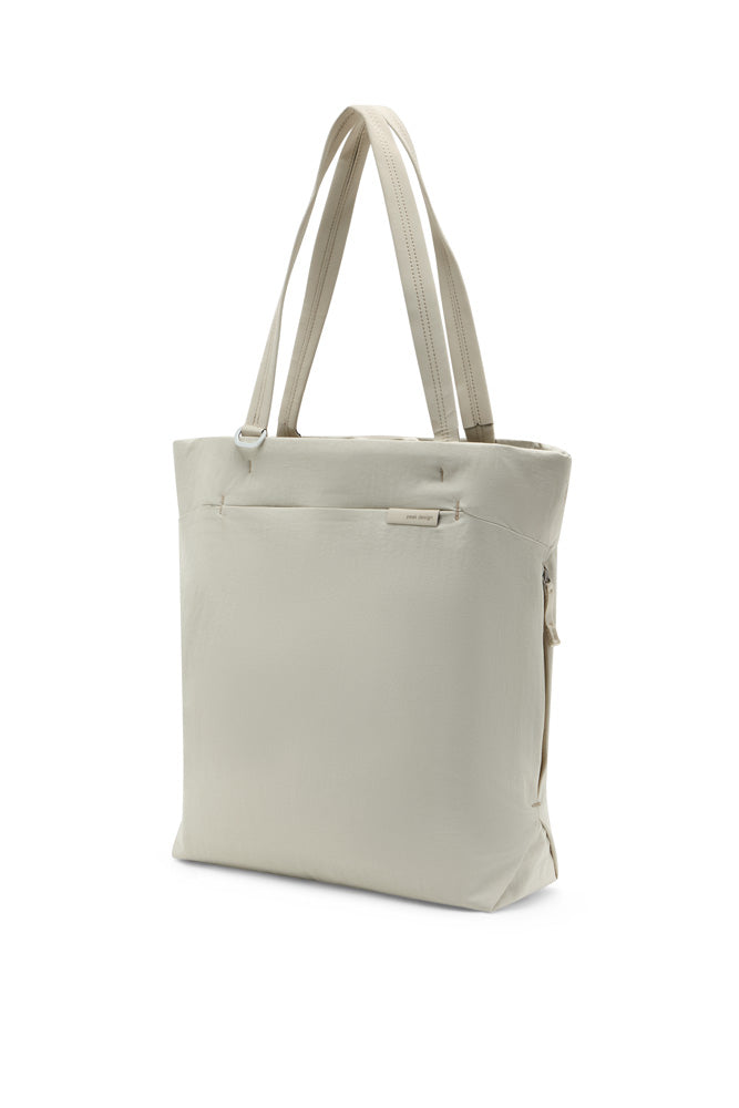 A light beige canvas tote bag with dual shoulder straps, shown from a three-quarter angle against a plain white background.