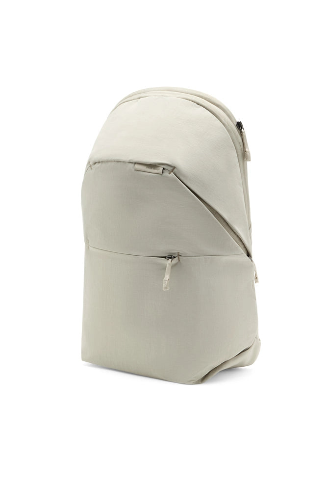 A light beige backpack with a curved top design, featuring a diagonal front zipper and a zippered pocket on the lower front, shown against a plain white background.