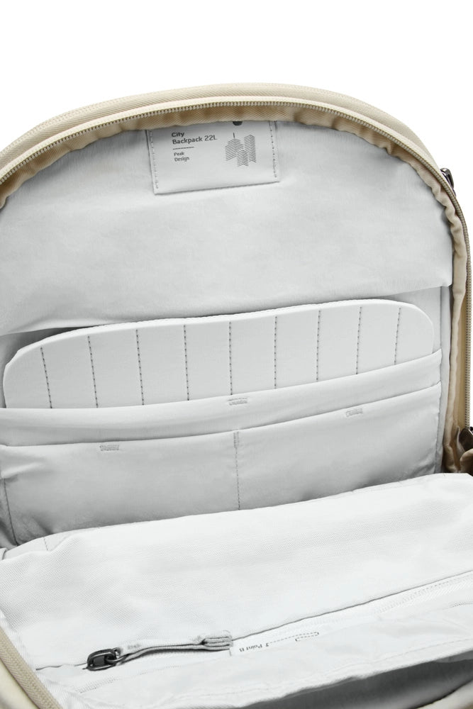 Close-up of the interior of a backpack showing a light gray padded compartment, zippered pockets, and a beige outer edge with a small sewn-in label.