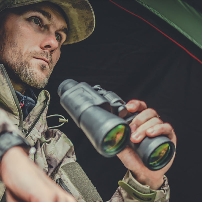A man in camouflage clothing inside a tent holds a pair of black binoculars up to his eye, looking through them.