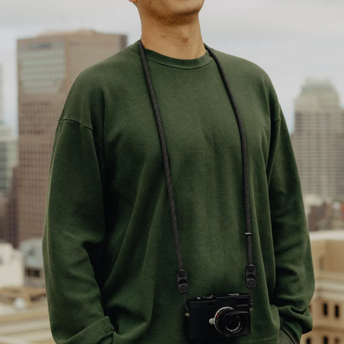 Person wearing a green sweatshirt with a camera around their neck, standing against a cityscape background.