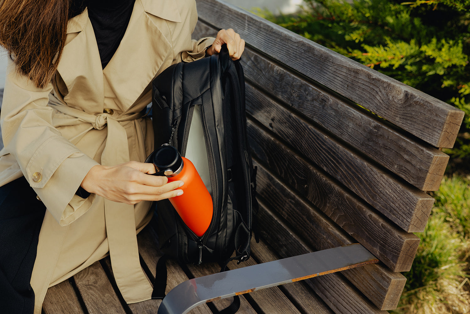 Person sitting on a wooden bench holding an orange water bottle and black backpack.
