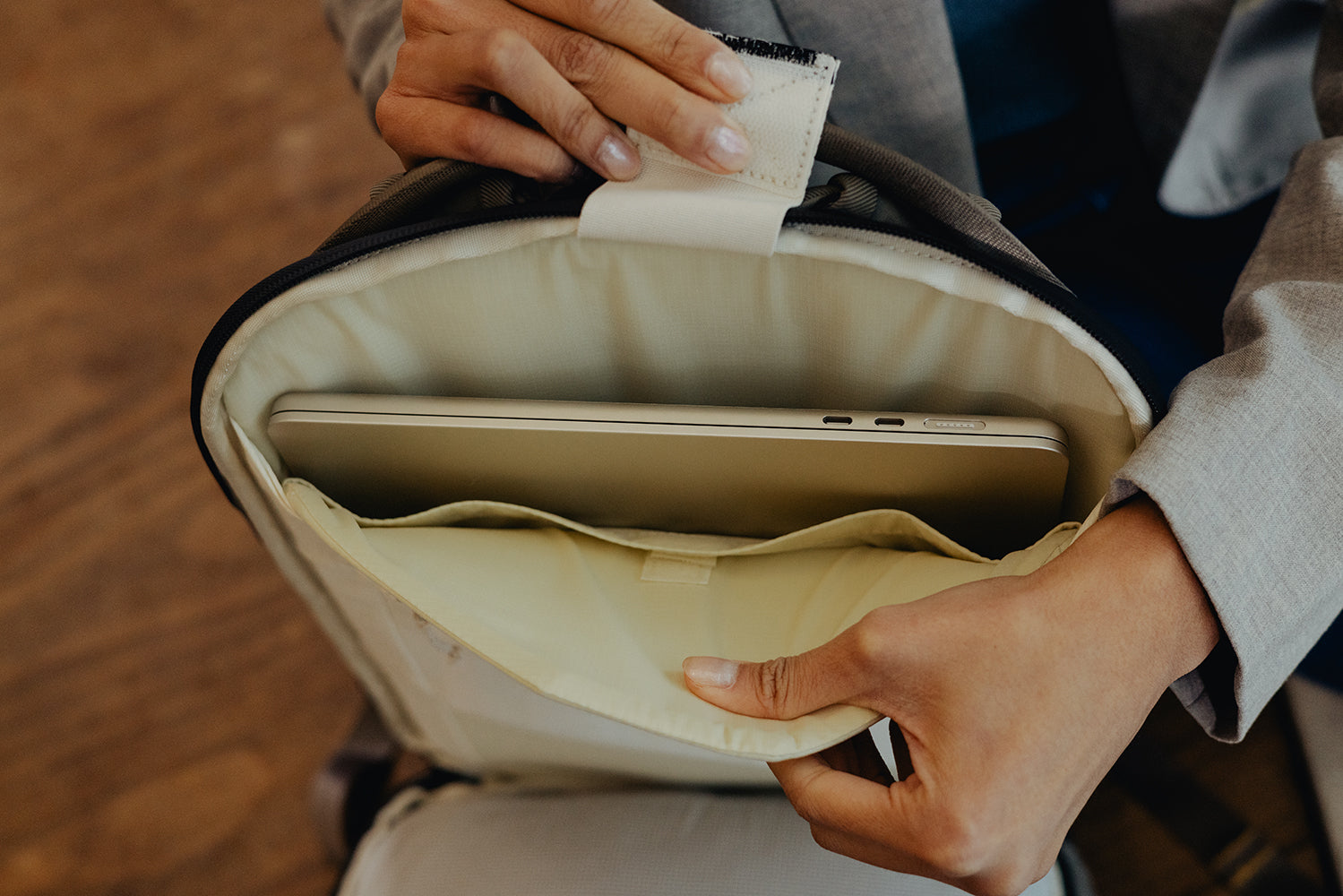 Person opening a beige backpack to reveal a laptop inside, on a wooden floor.